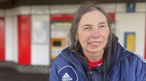 Paula Mitchinson is smiling against the red and white backdrop of the Stadium of Light metro station. She is wearing a blue Adidas puffer jacket with Sunderland AFC's crest and has short grey hair.