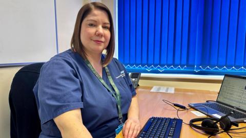 A nurse is sat at a desk with a headset and computer. She has a blue NHS top and lanyard and has short brown hair.