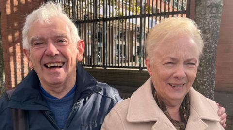 Alan and Joan Bell. Alan has white hair and is wearing a dark blue rain coat and blue jumper. Joan has short blonde hair and wearing a beige coat and brown scarf.