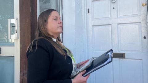 A woman with a clipboard and lanyard outside a white-painted wooden door.