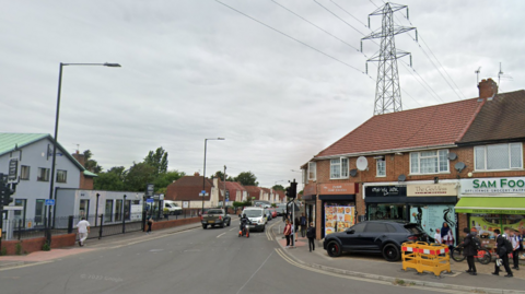 The road junction of Stoke Road and Elliman Avenue in Slough. It is a four-way junction with traffic lights. Along the road are a number of terraced houses.