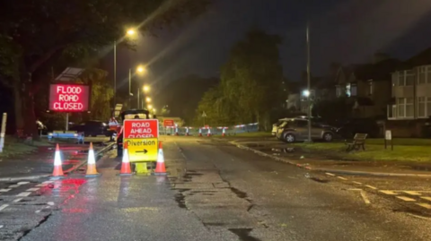 Shot of Queens Drive in Liverpool after a flood. A sign telling drivers 'Flood Road Closed' stands on the pavement with a diversion sign next to it. The picture was taken on a dark night.