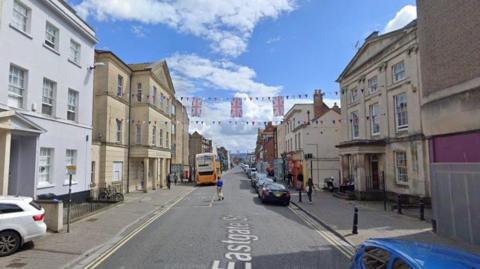 A streetview image of Eastgate Street in Gloucester, taken from the middle of the road. It is lined with old buildings built of light brick, and there are cars parked along the right-hand side. An orange bus is visible in the distance. 