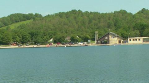 A wide outdoor scene with a large body of calm water in the foreground.
Across the water, there is a shoreline with several buildings and a small parking area. On the left side, there is a large modern building with a sloped roof and wide windows. To the right, there is a smaller building with a pitched roof and light-colored walls, and another low building beside it. A number of parked cars are lined up along the edge of the water, and a few small figures can be seen walking or standing near the shoreline. Behind the buildings, the background is filled with dense green trees covering rolling hills.