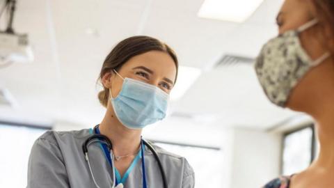 A doctor wearing a blue face mask while looking at a patient with a flowery mask on. They are sat inside a white hospital room.