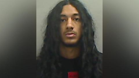 Mugshot of Mirza. He has very long thick black hair, a thin black beard and is wearing a black jumper with a red square logo beneath the neckline.