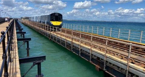 A picture of a Network Rail train, which has yellow detailing at the front and black panelling, is riding along a railway line, fixed above the sea on the Island Line. A pier runs alongside the line, decorated with black metal railings and people can be seen walking along the pier in the distance.