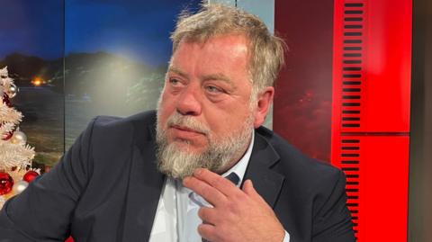 A man with his hand on his chin wearing a suit sitting in the BBC Channel Islands TV studio.