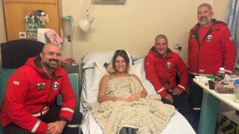Women in hospital bed with mountain rescue standing around her
