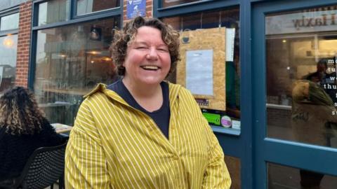 A lady is pictured smiling at the camera. She wears a bright yellow shirt with a navy t-shirt underneath.