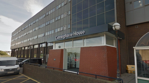 A large office building, with a rad brick entrance below many glass windows. Further along the building are rows of other windows.