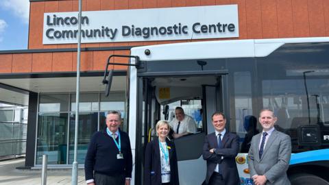 A wide angle shot of a bus outside Lincoln Community Diagnostic Centre. There are four people stood outside the bus. A driver is sat behind the steering wheel. 