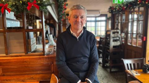 A man with greying hair and a dark blue shirt. He is sitting inside a pub.