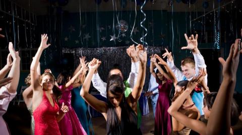 A stock photo of teenagers (15-18) in formalwear dancing at prom, arms raised. Streamer decorations hang from the ceiling.