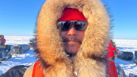 Barnie Aggark, an Inuk man, pictured wearing a hood with a thick fur trim and sunglasses. (for the photo captions). 
