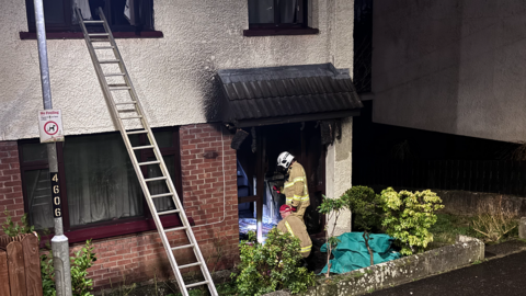 A home showing signs of smoke damage with two fire fighters standing in uniform at the door. 