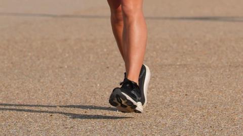 A close-up image of legs and black and white trainers running. The ground is a sandy colour.
