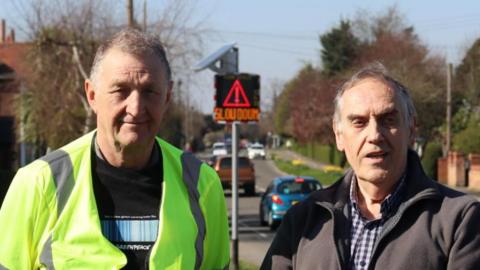 A view of two men stood in front of a speed indicator device, which is currently flashing a red caution sign urging a driver to slow down.