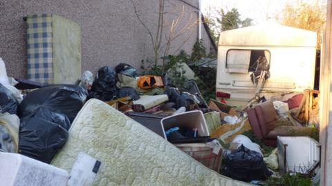 Piles of rubbish stacked up at the side of a property. The image shows an abandoned caravan to the back right of the picture. In the foreground are mattresses, bed bases, a sofa, black bin bags and various detritus. 