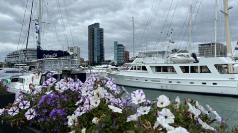 Purple and white flowers in a basket on railings alongside Ocean Village marina are in the foreground with several white boats behind. On the quayside are four large buildings. The day is overcast with grey skies.