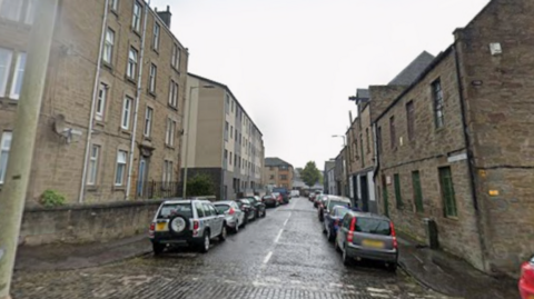 A cobbled street with tenements and cars on either side