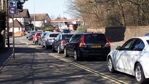 Traffic on the A460 Featherstone, with cars queueing on a built-up road with double yellow lines and a sign showing a 30mph speed limit and a speed camera warning. There are houses and a petrol station ahead.