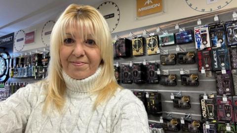 Jenny Phillips has long blonde hair and is wearing a white jumper. She is standing in one her sports gear shops with darts merchandise is on the wall behind her.