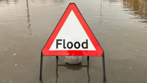 Red and white triangle flood warning sign standing in murky looking water. There is a sandbag at the rear of the sign holding it in place.