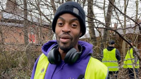 A man with a black beard wearing black headphones around his neck, a purple hoodie, a bright yellow hi-vis and a black beanie smiles at the camera. In the background are tree, bushes and other litter pickers.