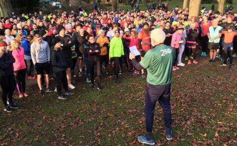 Large group of people in running gear listen to a man speaking before starting on a parkrun in Hull's East Park