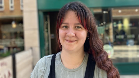 Sian has brown hair with red highlights on her fringe is wearing a grey blue top and black dungarees over it.  