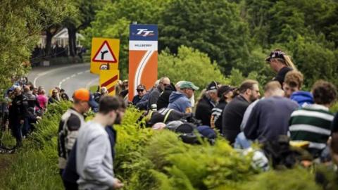 Spectators lining the hedge at Hillberry on the TT course. A sign for the milestone can be seen alongside other road signs, as the road sweeps up to the right.