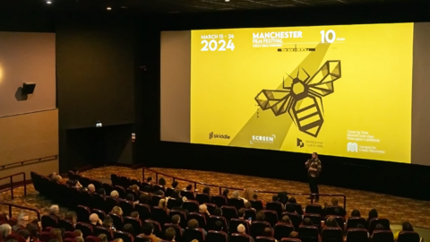 Yellow cinema screen showing a Manchester bee with a man holding a microphone in front of people sitting in seats waiting to watch a film.