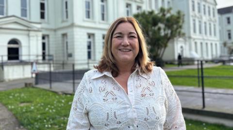 Jo Terry-Marchant wearing a white blouse. She has shoulder length brown hair and is stood in front of the white school.