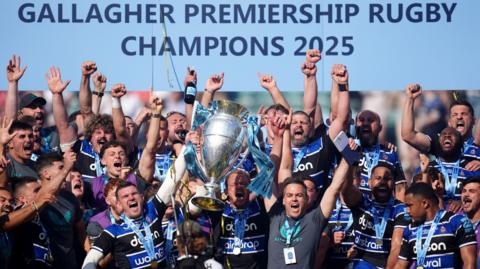 Bath captain Ben Spencer and head of rugby Johann van Graan hold the Premiership trophy in the air surrounded by the rest of the team as they celebrate on the podium at Twickenham