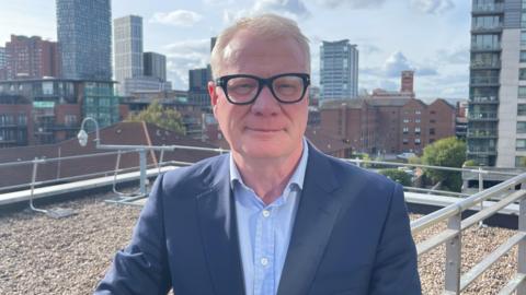 A man with grey hair and glasses who is wearing a dark blue suit and light blue tie. He is standing on a roof top with buildings in part of Birmingham visible behind him. 