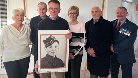 Group of six people posing for photo. Jerome Donnini is second from the right. There is also a man wearing a suit and medals on his breast-pocket. 
Two other women and three men, are dressed smartly. The man in the middle is holding a large photo of Dennis Donnini wearing a military hat. 