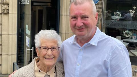 A man in a white shirt stands with his arm around a woman with short white hair and glasses and wearing a cream shirt. A building behind them.