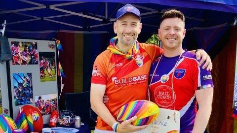 Two men, one standing with his arm around the other. They both have short hair and slight beards. One has a purple cap. One is wearing an orange T-shirt and the other a purple and red T-shirt. They are holding a rainbow-coloured rugby ball. They are under a blue gazebo with a metal frame and there is a noticeboard to their right with various photographs.