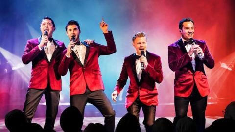 The Barricade Boys performing. All members are wearing red velvet suit jackets, bowties and black trousers. 