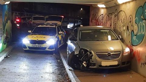 A silver Vauxhall that has crashed into the wall inside a tunnel. It's right wheel, fender and bonnet have been crumpled up. Behind it there is a police car parked with its light on, illuminating the road inside the tunnel. On the walls there are lots of graffiti tags.