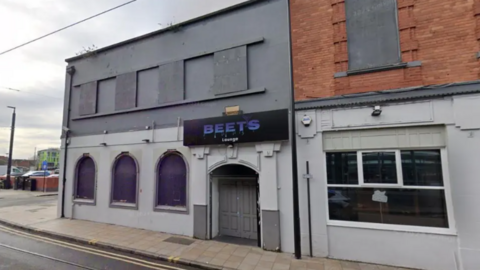 The building is painted grey with a purple/grey sign outside above the entrance. Upper and lower windows appear blocked out. It sits on the end of a street.