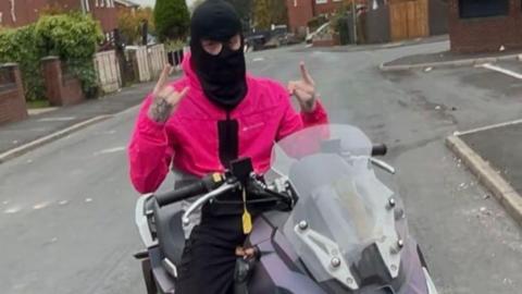 Teen poses on a bike wearing a balaclava making hand gestures