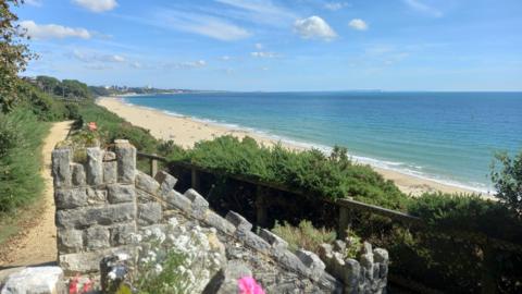 A scene of a beach from a cliff above. The sky is blue with just a few fair weather clouds. A stone structure - possibly steps - can be seen in the foreground with a tree line running behind the beach.