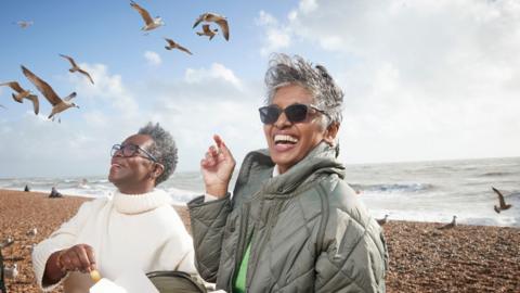 Two women smiling whilst eating fish and chips on a British pebble beach. Seagulls surround them and the sea is in the background.