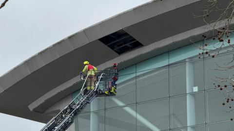 An emergency service worker is stood on an aerial platform as he investigates a missing rectangular sheet of metal from a shopping centre roof