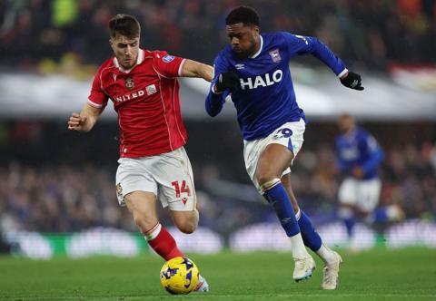 A Wrexham footballer in a red t-shirt and white shorts on the pitch with an Ipswich footballer in a blue t-shirt and white shorts. The ball is in play and is yellow