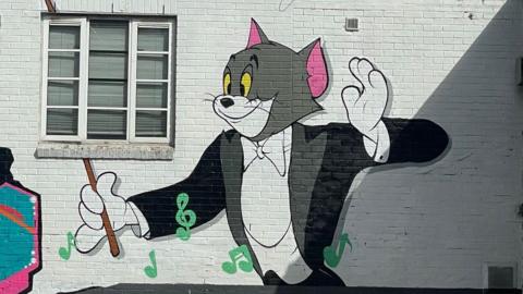 A street art mural featuring cartoon cat Tom, from Tom and Jerry, conducting music with green note symbols floating around,