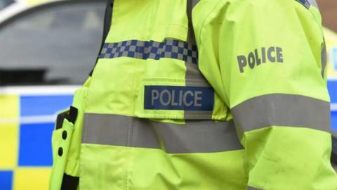 A close up of a police officer's jacket in hi-vis with the words 'police' written on it, and a police car in the background.