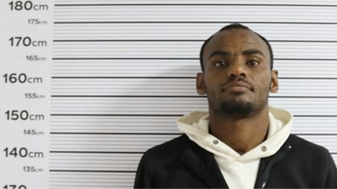 Yetbarek Dawit in a black top over a cream hoodie stands against a measuring chart in a police photo - putting his height at around 175cm.